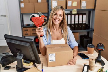 Young blonde woman ecommerce business worker holding packing tape machine at office