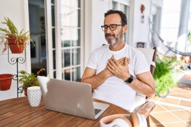 Middle age man using computer laptop at home smiling with hands on chest with closed eyes and grateful gesture on face. health concept. 