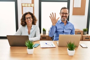 Middle age hispanic woman and man sitting with laptop at the office showing and pointing up with fingers number five while smiling confident and happy. 