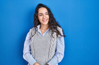 Mavi arka planda duran genç esmer kadın yüzünde gülümseme, doğal bir ifade ile yan yana bakıyor. Kendinden emin gülen. 