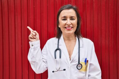 Middle age hispanic woman wearing doctor uniform and stethoscope smiling happy pointing with hand and finger 
