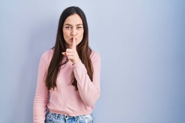 Genç esmer kadın mavi arka planda dikiliyor ve parmakları dudaklarında sessiz olmak istiyor. sessizlik ve gizli konsept. 