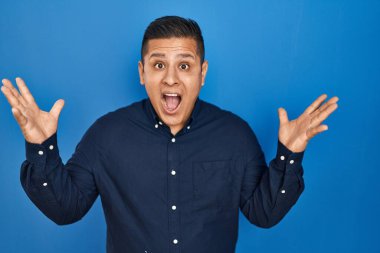 Hispanic young man standing over blue background celebrating crazy and amazed for success with arms raised and open eyes screaming excited. winner concept 