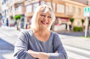 Orta yaşlı sarışın kadın gülümsüyor. Kollarını kavuşturup caddede duruyor.