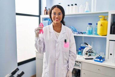 Genç İspanyol doktor, elinde pembe kurdeleyle laboratuvarda çalışıyor. Pozitif görünüyor ve gülümsüyor. Kendine güvenen bir gülümsemeyle dişlerini gösteriyor. 