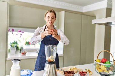 Genç sarışın kadın mutfaktaki blendere muz dökerek gülümsüyor.