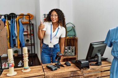 Giyim mağazasında çalışan güzel İspanyol kadın mutlu ve olumlu gülümsüyor, başparmağıyla mükemmel ve onay işareti yapıyor. 