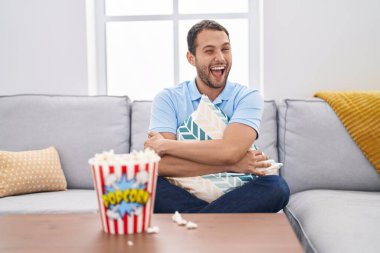 Hispanic man watching tv at home sitting on the sofa winking looking at the camera with sexy expression, cheerful and happy face. 