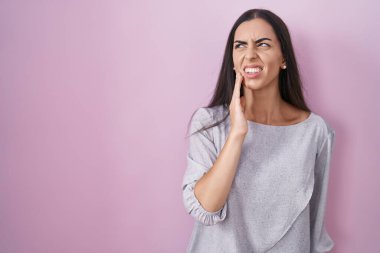 Young brunette woman standing over pink background touching mouth with hand with painful expression because of toothache or dental illness on teeth. dentist 