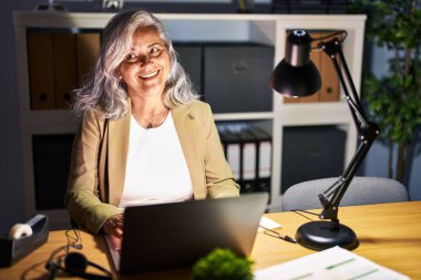 Middle age woman with grey hair working using computer laptop late at night smiling looking to the side and staring away thinking. 