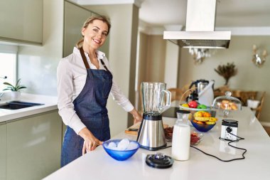 Genç sarışın kadın gülümsüyor. Mutfakta kendine güvenen smoothie pişiriyor.