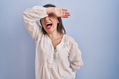 Middle age hispanic woman standing over blue background covering eyes with arm smiling cheerful and funny. blind concept. 