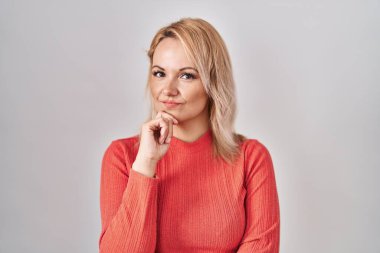 Sarışın kadın izole arka planda duruyor kameraya bakıyor kolları çapraz ve elleri çenesinde gülümsüyor. Olumlu düşünmek. 