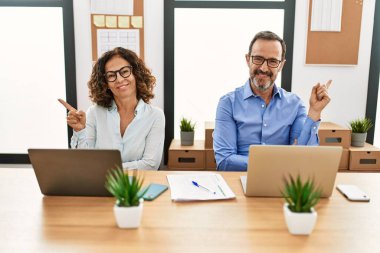 Middle age hispanic woman and man sitting with laptop at the office with a big smile on face, pointing with hand and finger to the side looking at the camera. 