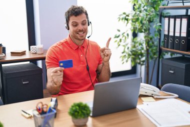 Çağrı merkezi kulaklığı takan genç İspanyol adam elinde kredi kartıyla gülümsüyor. 