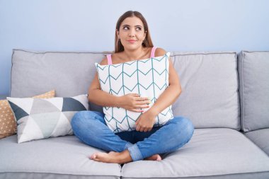 Young blonde woman hugging pillow sitting on the sofa smiling looking to the side and staring away thinking. 
