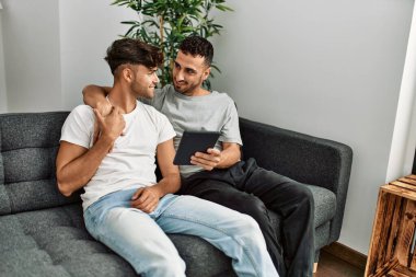 Two hispanic men couple using touchpad hugging each other at home