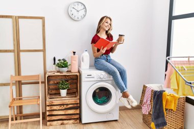 Young caucasian girl drinking coffee and reading book waiting for laundry sitting on whasing machine at home.