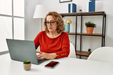 Orta yaşlı beyaz kadın evde dizüstü bilgisayar kullanıyor. Tavırlarını ve ayık ifadesini düşünüyor. Kendine güveniyor gibi görünüyor. 