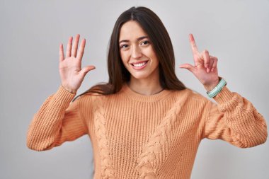 Genç esmer kadın beyaz arka planda duruyor. Yedi numaralı parmaklarıyla işaret ediyor ve gülümsüyor. Kendinden emin ve mutlu.. 