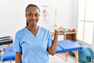 Saçları örgülü siyah bir kadın, ağrı giderme kliniğinde çalışıyor. Gülümsüyor, gülümsüyor ve avuç içiyle kameraya bakıyor.. 