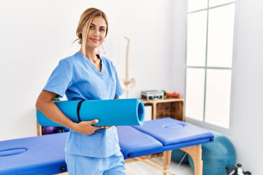 Kafkasyalı genç fizyoterapist klinikte yoga minderi tutarken mutlu gülümsüyor.