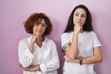 İspanyol anne ve kızı pembe arka plan üzerine günlük beyaz t-shirt giyiyorlar ve elleri çenelerinde soru sorup, düşünceli bir ifade takınıyorlar. Düşünceli bir yüzle gülümsüyor. şüphe kavramı. 