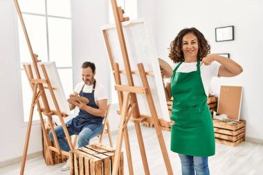 Hispanic middle age woman and mature man at art studio pointing finger to one self smiling happy and proud 