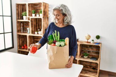 Orta yaşlı, gri saçlı, elinde poşetlerle evde bekleyen bir kadın..