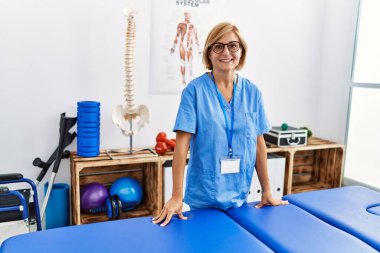 Klinikte fizyoterapist üniforması giyen orta yaşlı sarışın kadın.