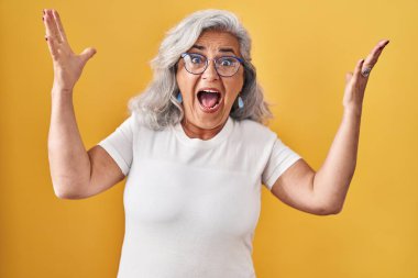 Middle age woman with grey hair standing over yellow background celebrating crazy and amazed for success with arms raised and open eyes screaming excited. winner concept 