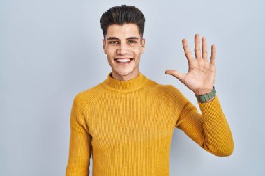 Mavi arka planda duran genç İspanyol adam beş numarayı gösterip işaret ederken kendinden emin ve mutlu gülümsüyor.. 