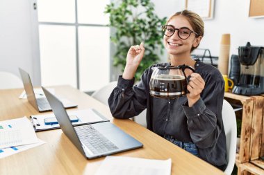 Ofiste çalışan genç sarışın kadın elinde kahve fincanıyla mutlu bir şekilde gülümsüyor. 