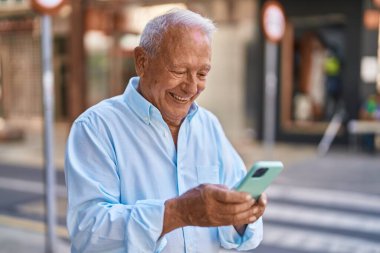 Yaşlı, gri saçlı, kendine güvenen, caddede akıllı telefon kullanan bir adam.