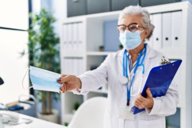 Doktor üniforması giyen yaşlı gri saçlı kadın klinikte tıbbi maske takıyor.