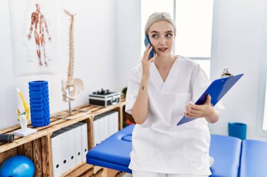 Fizyoterapist üniforması giyen beyaz bir kadın klinikte akıllı telefondan konuşuyor.