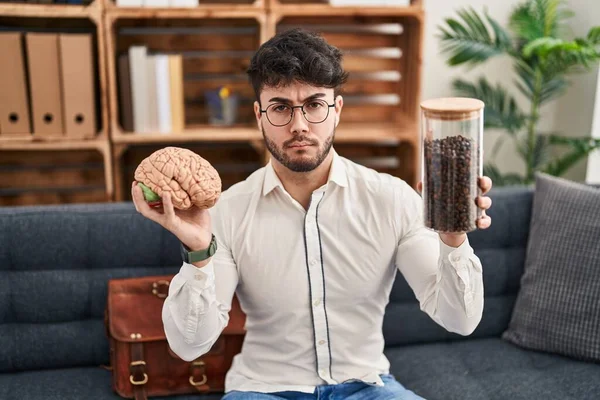 Hispanic man with beard working at therapy office holding brain and coffee beans depressed and worry for distress, crying angry and afraid. sad expression.