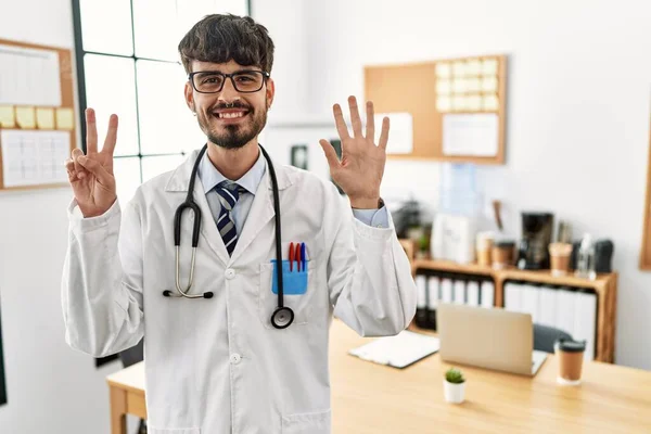 在办公室里 留着胡子 身穿医生制服和听诊器的西班牙裔男子露出七号手指 面带微笑 自信而快乐 — 图库照片