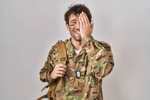 顔に自信のある笑顔と驚きの感情で一つの目を覆うカモフラージュ軍の制服を着たヒスパニック系の若者 — ストック写真
