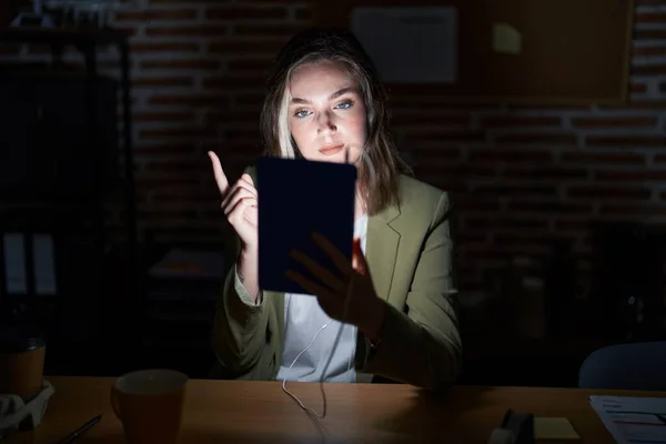 Blonde caucasian woman working at the office at night pointing with hand finger to the side showing advertisement, serious and calm face