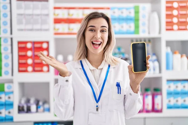 Young beautiful woman working at pharmacy drugstore showing smartphone screen celebrating achievement with happy smile and winner expression with raised hand
