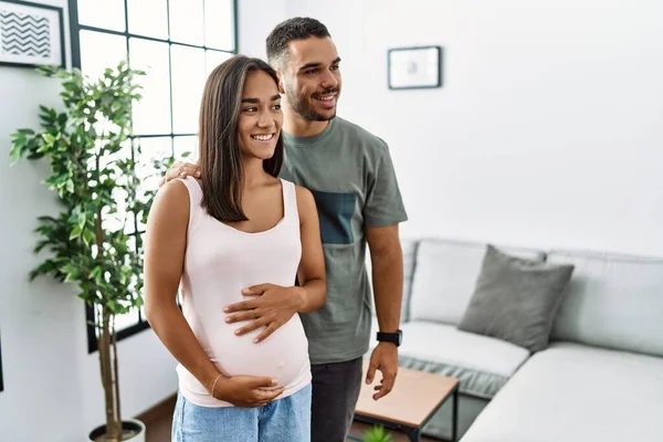 Young interracial couple expecting a baby, touching pregnant belly looking away to side with smile on face, natural expression. laughing confident.