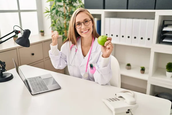 年轻的金发女医生拿着青苹果站在诊所 手指指向旁边 笑容满面 张开嘴 — 图库照片