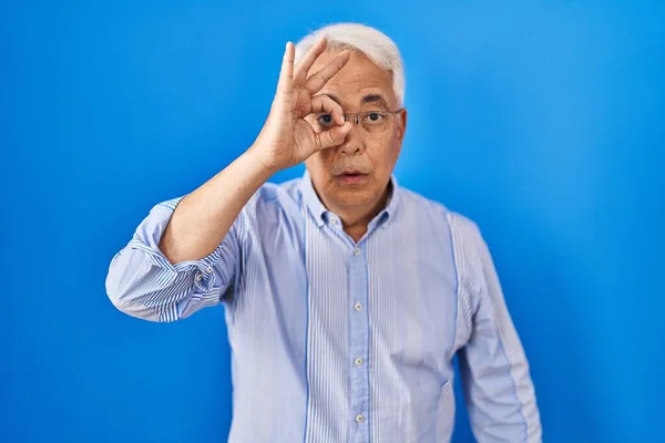 Hispanic senior man wearing glasses doing ok gesture shocked with surprised face, eye looking through fingers. unbelieving expression.