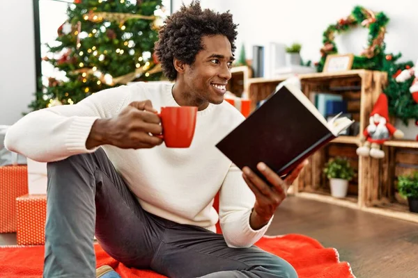 若いですアフリカ系アメリカ人男性読書本とコーヒーを飲みます床の上に座っていますクリスマスツリーによってホーム — ストック写真