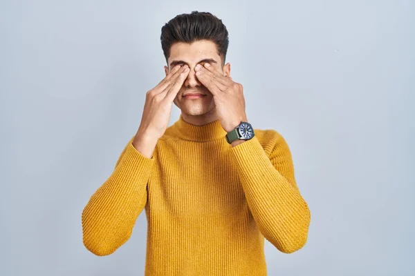 Young hispanic man standing over blue background rubbing eyes for fatigue and headache, sleepy and tired expression. vision problem
