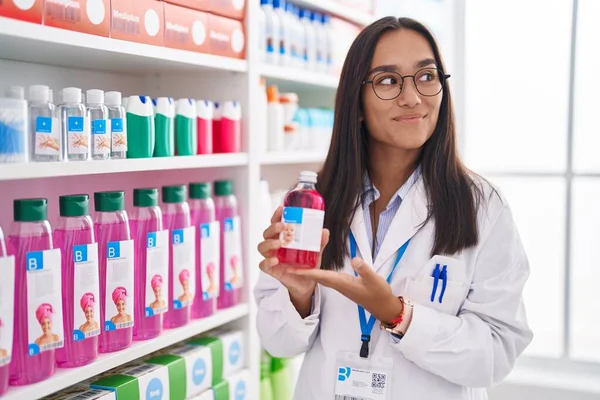 Young hispanic woman working at pharmacy drugstore holding syrup smiling looking to the side and staring away thinking.