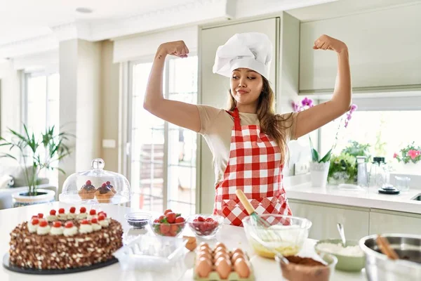 Beautiful young brunette pastry chef woman cooking pastries at the kitchen showing arms muscles smiling proud. fitness concept.