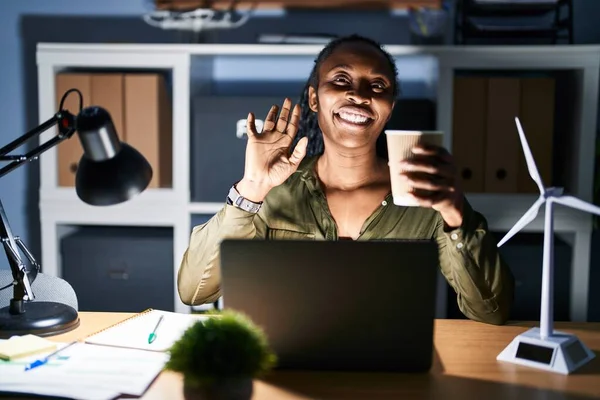 African woman working using computer laptop at night waiving saying hello happy and smiling, friendly welcome gesture