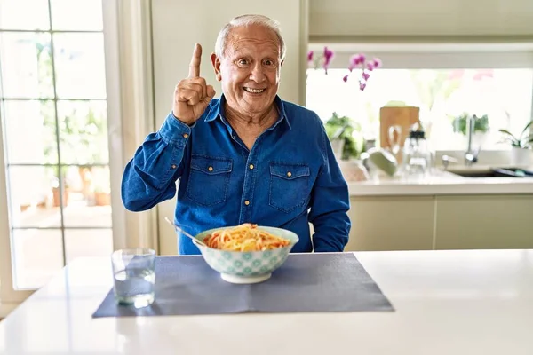 Senior man with grey hair eating pasta spaghetti at home pointing finger up with successful idea. exited and happy. number one.